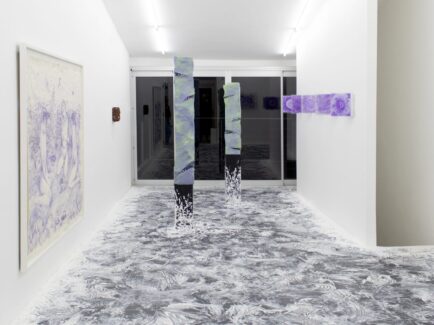 Una sala blanca con obras de arte, el piso gris está manchado, hay cuadros color violeta