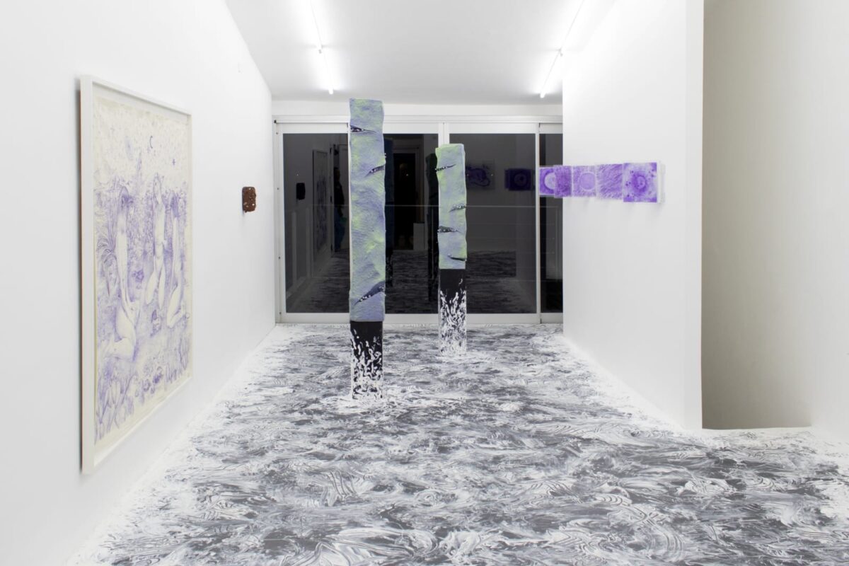 Una sala blanca con obras de arte, el piso gris está manchado, hay cuadros color violeta